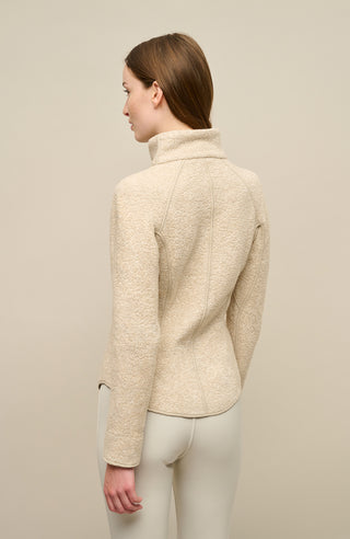 Ein Model trägt die Damenjacke „WERA-CT 94-0“ von Frauenschuh in der Farbe „Caramel“, einem hellen, warmen Beigeton. Die Jacke besteht aus luxuriösem Teddyfleece mit Kaschmiranteil, das sich durch seine weiche, voluminöse Struktur auszeichnet. In der Rückansicht sichtbar ist der hohe Stehkragen sowie der leicht gerundete Saum, der seitlich höher geschnitten ist und dem Modell eine moderne, dynamische Passform verleiht.