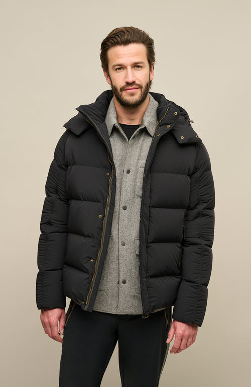 Vorderansicht der Frauenschuh Daunenjacke Mattia für Herren in Schwarz die von einem Model getragen wird – warme Winterjacke mit abknöpfbarer und verstellbarer Kapuze, modernem Steppmuster, verschließbaren Seitentaschen und zusätzlicher Knopfleiste. Sie ist außerdem Feuchtigkeitsregulierend, Wasserabweisend, Wärmend, Atmungsaktiv und Elastisch.