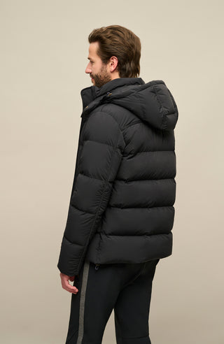 Rückansicht der Frauenschuh Daunenjacke Mattia für Herren in Schwarz die von einem Model getragen wird – warme Winterjacke mit abknöpfbarer und verstellbarer Kapuze, modernem Steppmuster, verschließbaren Seitentaschen und zusätzlicher Knopfleiste. Sie ist außerdem Feuchtigkeitsregulierend, Wasserabweisend, Wärmend, Atmungsaktiv und Elastisch.