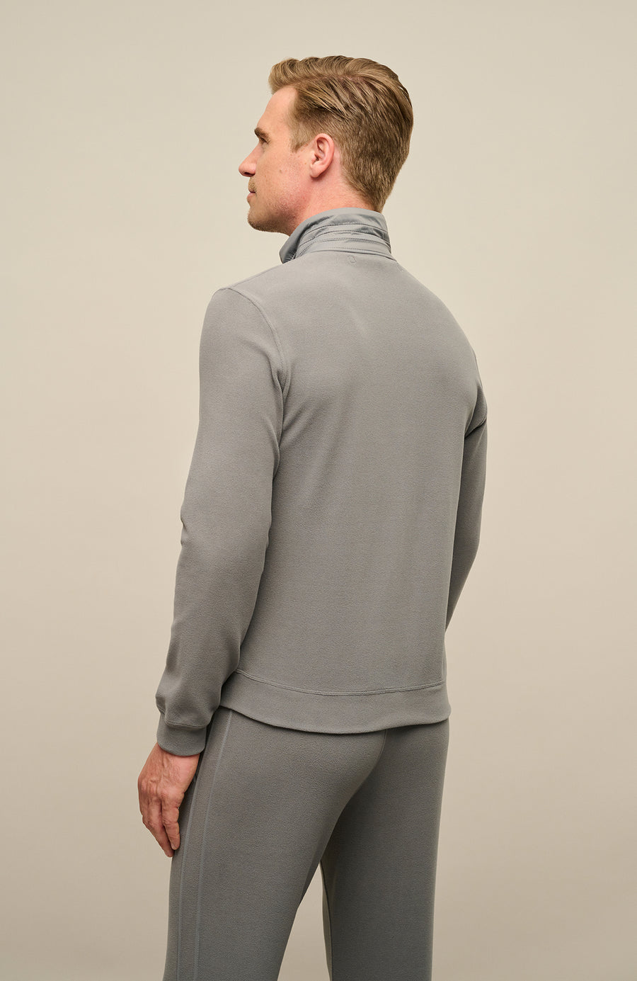 Rückansicht der Herrenjacke „Oliver-BF 2520“ von Frauenschuh in der Farbe „Cement“, einem hellen, modernen Grauton mit leicht warmem Unterton. Die Jacke wird von einem Model getragen und zeigt eine klare, reduzierte Rückseite mit dezenter Nahtführung und ergonomisch geformtem Schnitt. Der hohe Stehkragen und die glatte, leicht strukturierte Materialoberfläche unterstreichen den funktionalen, stilvollen Charakter des Designs. Die leicht taillierte Silhouette sorgt für eine sportlich-elegante Optik