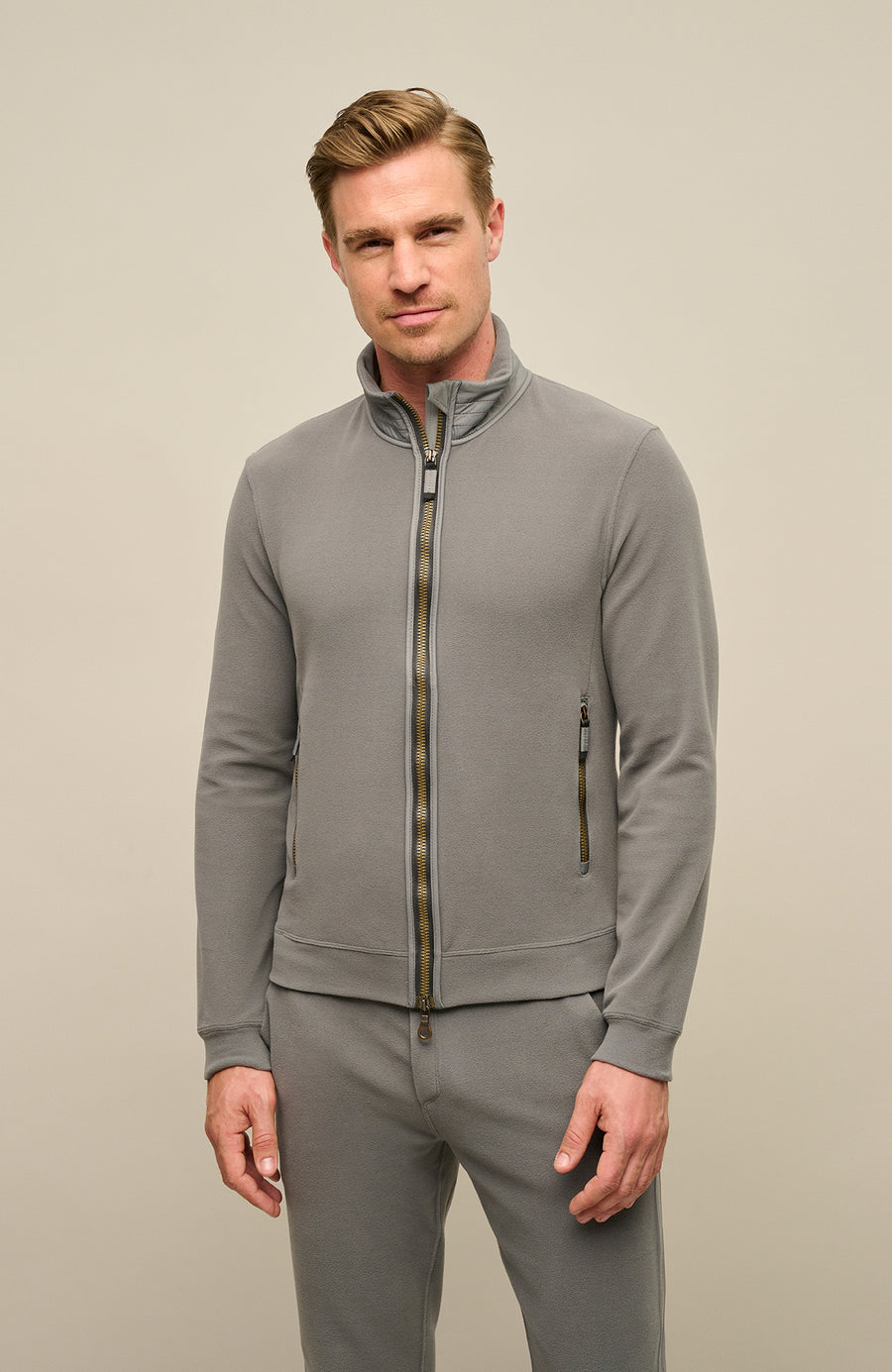 Herrenjacke „Oliver-BF 2520“ in der Farbe „Cement“ – ein heller, neutraler Grauton mit leicht warmem Unterton. Die Jacke ist in der Vorderansicht abgebildet, wird von einem Model getragen und präsentiert sich mit klarem, sportlich-elegantem Design. Sie verfügt über einen durchgehenden Reißverschluss, einen hohen Stehkragen sowie zwei seitlich platzierte Reißverschlusstaschen. Das Material wirkt leicht, funktional und strukturiert 