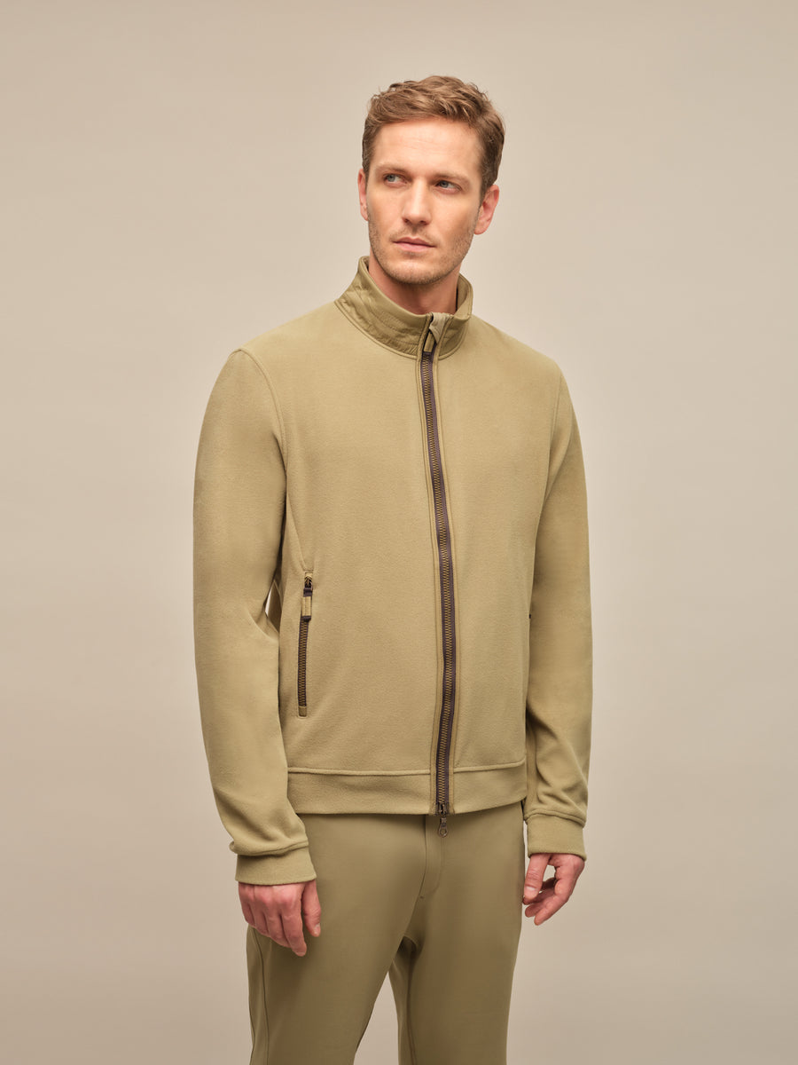 Ein männliches Model trägt eine modische, leichte Herrenjacke in der Farbe "Sage" (ein dezentes, graugrünes Pastell), die Teil der Frühjahrs- und Sommerkollektion von Frauenschuh Mode ist. Die Jacke hat eine moderne, sportlich-elegante Passform mit einem hohen Kragen und einem durchgehenden Reißverschluss vorne. Dezente Nahtdetails und funktionale Taschen mit minimalistischen Reißverschlüssen unterstreichen das hochwertige Design. Die Ärmel sind leicht vorgeformt und mit dezenten Bündchenabschlüssen versehe