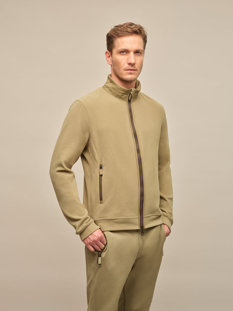 Ein männliches Model trägt eine modische, leichte Herrenjacke in der Farbe "Sage" (ein dezentes, graugrünes Pastell), die Teil der Frühjahrs- und Sommerkollektion von Frauenschuh Mode ist. Die Jacke hat eine moderne, sportlich-elegante Passform mit einem hohen Kragen und einem durchgehenden Reißverschluss vorne. Dezente Nahtdetails und funktionale Taschen mit minimalistischen Reißverschlüssen unterstreichen das hochwertige Design. Die Ärmel sind leicht vorgeformt und mit dezenten Bündchenabschlüssen versehe