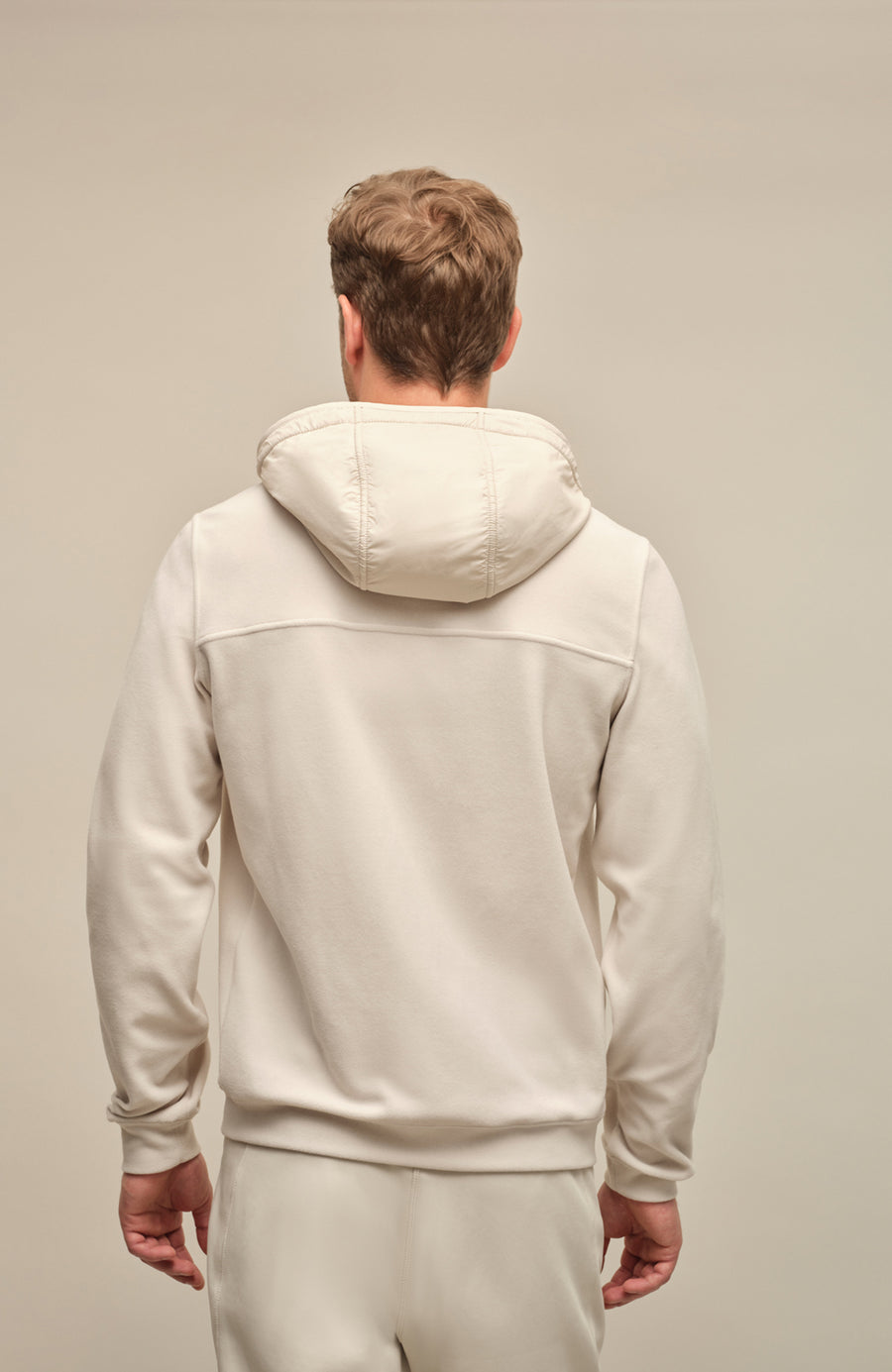 Rückansicht der Herrenjacke "Thomas" von einem Model getragen, in der Farbe Sandstone. Die Jacke zeichnet sich durch einen minimalistischen Schnitt, einen verstellbaren Nylon-Kapuzenkragen und praktische Reißverschlusstaschen aus. Ideal für alle Jahreszeiten, kombiniert sie Funktionalität mit modernem Design