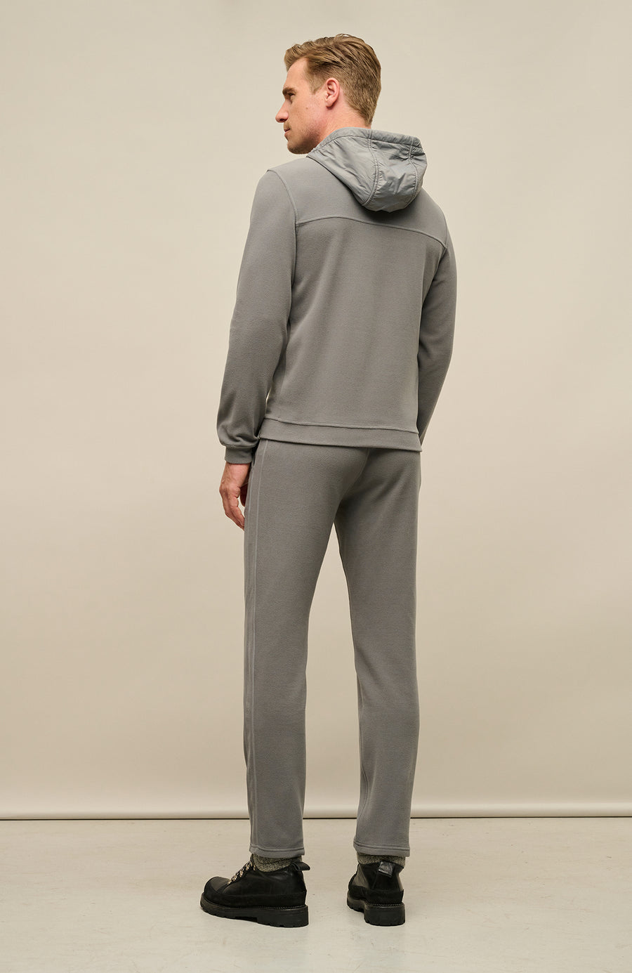 Rückansicht der Herrenjacke "Thomas" von einem Model getragen, in der Farbe grau. Die Jacke zeichnet sich durch einen minimalistischen Schnitt, einen verstellbaren Nylon-Kapuzenkragen und praktische Reißverschlusstaschen aus. Ideal für alle Jahreszeiten, kombiniert sie Funktionalität mit modernem Design