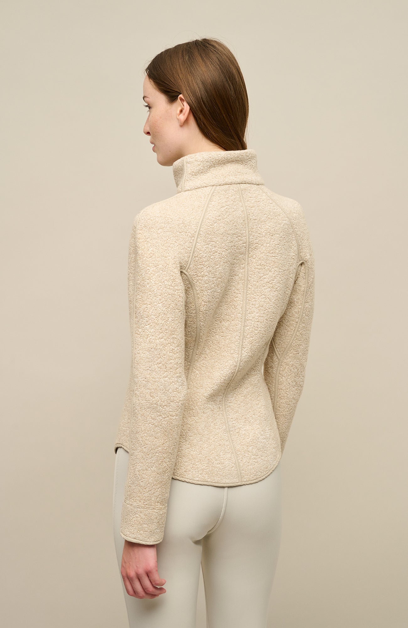 Ein Model trägt die Damenjacke „WERA-CT 94-0“ von Frauenschuh in der Farbe „Caramel“, einem hellen, warmen Beigeton. Die Jacke besteht aus luxuriösem Teddyfleece mit Kaschmiranteil, das sich durch seine weiche, voluminöse Struktur auszeichnet. In der Rückansicht sichtbar ist der hohe Stehkragen sowie der leicht gerundete Saum, der seitlich höher geschnitten ist und dem Modell eine moderne, dynamische Passform verleiht.