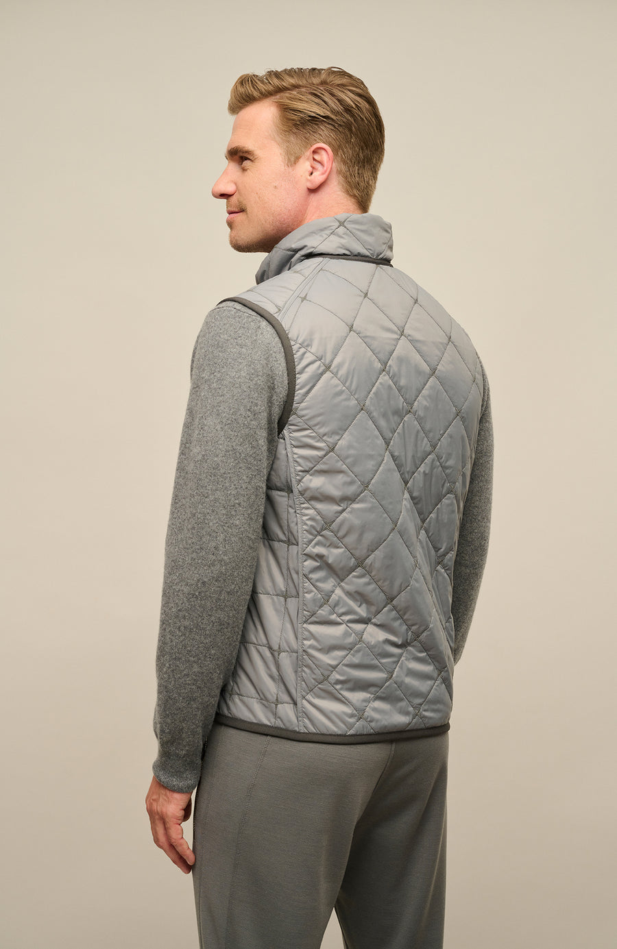 Rückansicht eines Herrenmodels mit der gesteppten Weste „Henri-QL“ von Frauenschuh in Cement (Grau); hochwertige Nylon-Qualität mit feiner Rautensteppung, sichtbar über die gesamte Rückenfläche; sportlich-taillierte Passform betont Schultern und Rücken; dezente Nahtführung für ein klares, modernes Design; ideal für die Übergangszeit Frühling und Sommer; das Model steht mit dem Rücken zur Kamera und trägt die Weste über einem leichten Oberteil – stilvolle Kombination aus Funktionalität und Luxusmode