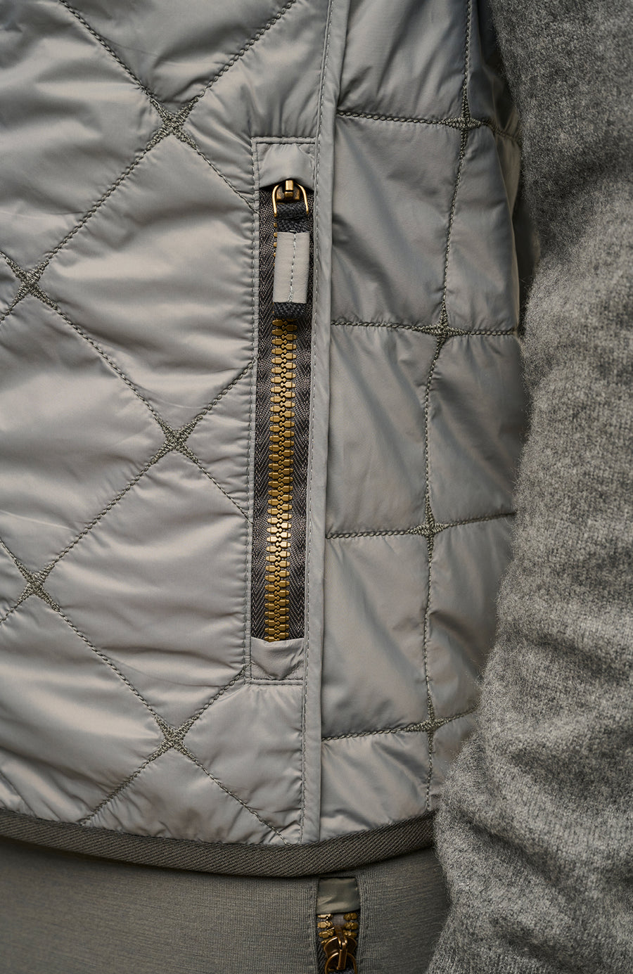 Detailansicht der Herren-Quiltweste „Henri-QL“ von Frauenschuh in der Farbe Cement (Grau); Fokus auf die seitliche Eingrifftasche mit hochwertigem Reißverschluss und integrierter Zipper-Garage – die den Zipper schützt und das minimalistische Design unterstreicht; sichtbar ist auch die feine Rautensteppung und die sorgfältige Verarbeitung des technischen Nylonmaterials; ideal für Frühling und Sommer als elegante, durchdachte Outdoor-Bekleidung