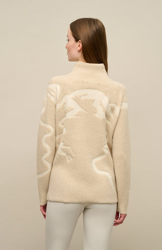 Modelrückansicht eines Damenpullovers aus 100 % extrafeiner Merinowolle, Modell „Chatel-ME“ in der Farbe Beige. Die Vorderseite zeigt ein kunstvoll gestaltetes Jacquard-Motiv aus skizzierten Berglandschaften, urbanen Elementen und dynamischen Skifahrern. Das hochwertige Merinogarn – leicht, weich und angenehm auf der Haut – wird in Italien produziert und auf modernsten Maschinen verarbeitet. Die Kombination aus künstlerischem Design & technischer Strickkunst macht den Pullover besonders stilvoll