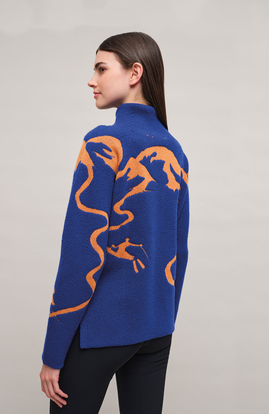 Rueckansiht am Model eines Damenpullovers aus 100 % extrafeiner Merinowolle, Modell „Chatel-ME“ in der Farbe Marineblau. Die Vorderseite zeigt ein kunstvoll gestaltetes Jacquard-Motiv aus skizzierten Berglandschaften, urbanen Elementen und dynamischen Skifahrern. Das hochwertige Merinogarn – leicht, weich und angenehm auf der Haut – wird in Italien produziert und auf modernsten Maschinen verarbeitet. Die Kombination aus künstlerischem Design & technischer Strickkunst macht den Pullover besonders stilvoll